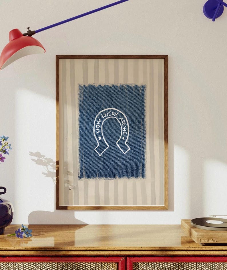 May include: Framed artwork featuring a blue denim-textured square with a white horseshoe design and the text "HOW LUCKY ARE WE" in a circular arrangement. The artwork is set against a striped background and framed in a wooden frame.