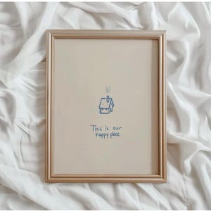 Peut inclure: Un cadre doré contenant une impression avec un fond beige clair. Un dessin au trait bleu d'une maison avec de la fumée s'élevant de la cheminée est centré au-dessus du texte bleu "This is our happy place".