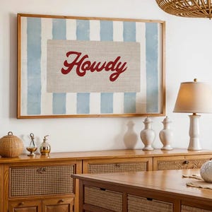 May include: Framed wall art featuring a light blue and white striped backdrop and a canvas with the word "Howdy" in red script. The artwork is displayed above a wooden cabinet.