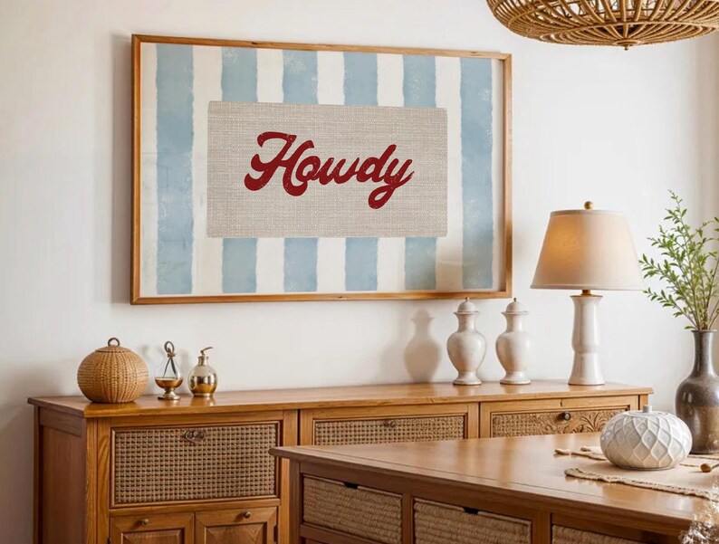 May include: Framed wall art featuring a light blue and white striped backdrop and a canvas with the word "Howdy" in red script. The artwork is displayed above a wooden cabinet.