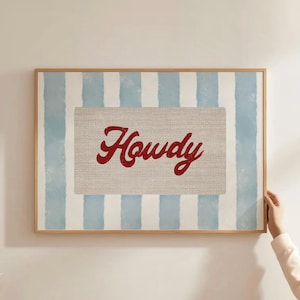 May include: Framed wall art with a light blue and white striped background. The centre features a rectangular canvas with the word "Howdy" in red script. The frame is made of light-coloured wood.