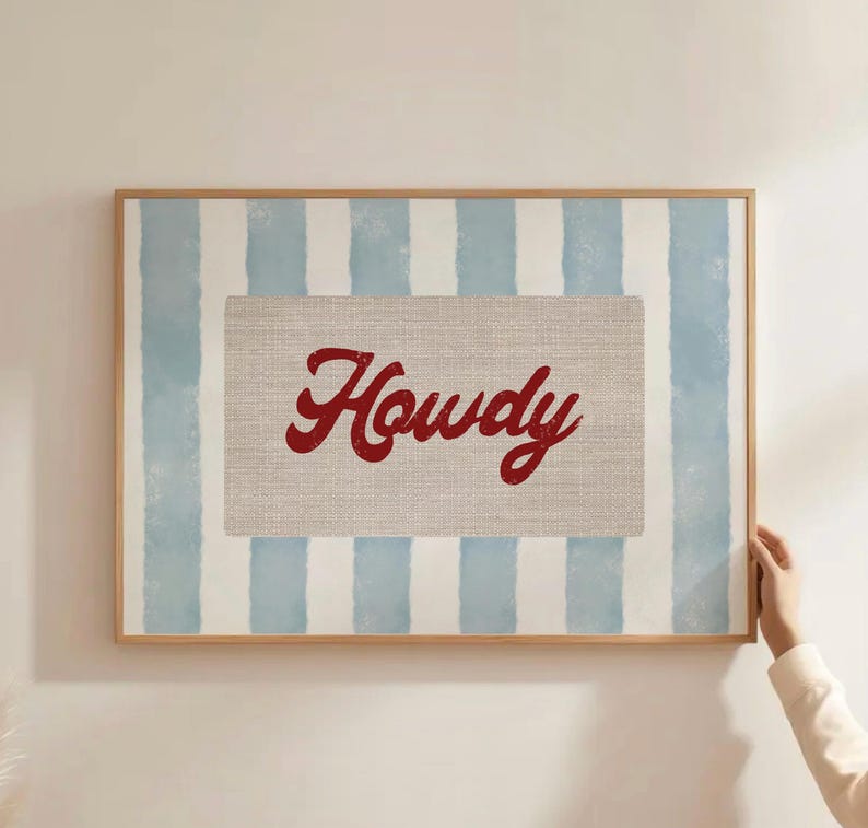 May include: Framed wall art with a light blue and white striped background. The centre features a rectangular canvas with the word "Howdy" in red script. The frame is made of light-coloured wood.