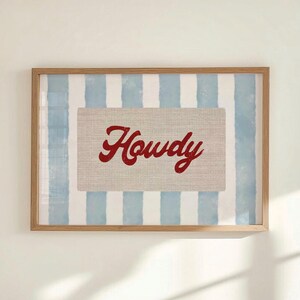 May include: Framed artwork with the word "Howdy" in red script on a beige rectangle. The background has blue and white vertical stripes, set within a light brown wooden frame.