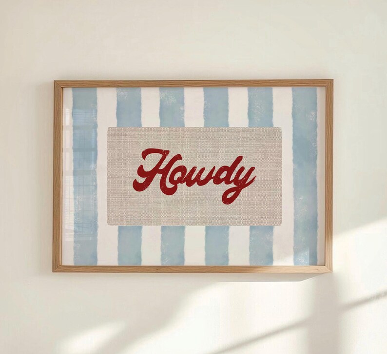 May include: Framed artwork with the word "Howdy" in red script on a beige rectangle. The background has blue and white vertical stripes, set within a light brown wooden frame.