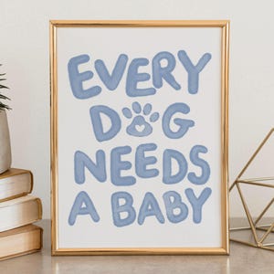Op de afbeelding: Een ingelijste print met de tekst "EVERY DOG NEEDS A BABY" in lichtblauw, met een pootafdruk en een hartje. De print zit in een gouden lijst en wordt op een neutraal oppervlak weergegeven. Een kleine plant in een pot en boeken zijn zichtbaar.