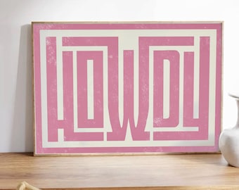 May include: A framed art print with the word "HOWDY" in a pink, block-style font. The word is centered on a cream-colored background, framed by a thin white border and a wider pink border. The print is displayed on a wooden surface.