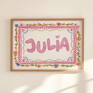 Może przedstawiać: Oprawiony wydruk akwarelowy z imieniem "JULIA" w r&oacute;żowych literach bąbelkowych. Imię jest umieszczone w białym prostokącie, otoczone ozdobną obw&oacute;dką z r&oacute;żowych, zielonych i kwiatowych akcent&oacute;w, wszystko w naturalnej drewnianej ramie.