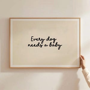 Op de afbeelding: Een ingelijste print met de tekst "Every dog needs a baby" in zwart schrift. De print heeft een crèmekleurige achtergrond en een lichtbruine houten lijst. Een hand houdt het frame vast.