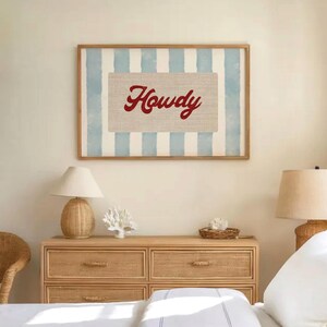 May include: A framed print featuring the word "Howdy" in red script on a beige background, set against a backdrop of light blue and white vertical stripes. The artwork is in a wooden frame and hangs above a wooden chest of drawers.