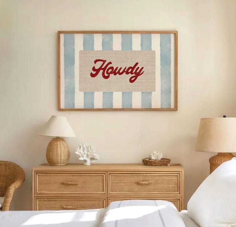 May include: A framed print featuring the word "Howdy" in red script on a beige background, set against a backdrop of light blue and white vertical stripes. The artwork is in a wooden frame and hangs above a wooden chest of drawers.