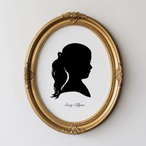 May include: A black silhouette of a young person with a ponytail, framed in an ornate gold oval frame. The silhouette is set against a white background. The name "Suzy Ellysa" is written in cursive below the silhouette.