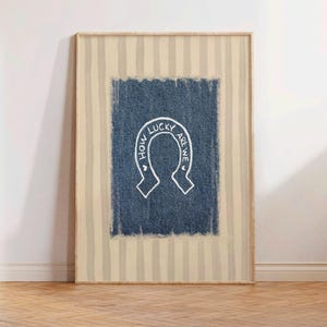 May include: Framed artwork featuring a denim-textured blue rectangle with a white horseshoe design. The horseshoe contains the text "HOW LUCKY ARE WE" in a handwritten style. The background has vertical beige and white stripes.