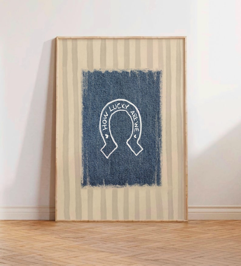 May include: Framed artwork featuring a denim-textured blue rectangle with a white horseshoe design. The horseshoe contains the text "HOW LUCKY ARE WE" in a handwritten style. The background has vertical beige and white stripes.