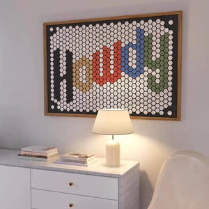 May include: A framed artwork spelling "HOME" in colourful hexagonal tiles. The art hangs above a white cabinet with a lamp and books. The lamp has a white base and a cream-coloured lampshade. The cabinet has brass knobs.