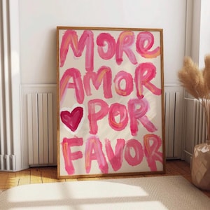 May include: A framed art print with the words "MORE AMOR POR FAVOR" in pink brushstrokes, with a pink heart. The artwork has a beige background and a wooden frame. The print is displayed on a wooden floor.