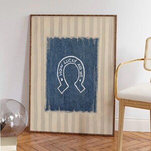 May include: Framed wall art featuring a blue denim-textured background with a white horseshoe design. The text "HOW LUCKY ARE WE" is written in a circular pattern around the horseshoe. The frame has a striped beige and white pattern.
