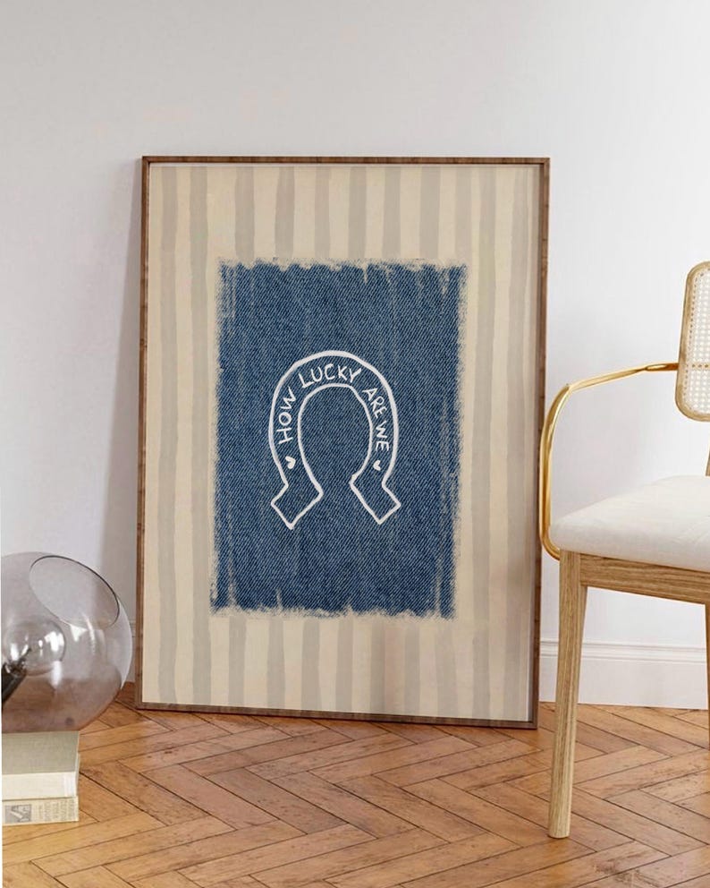 May include: Framed wall art featuring a blue denim-textured background with a white horseshoe design. The text "HOW LUCKY ARE WE" is written in a circular pattern around the horseshoe. The frame has a striped beige and white pattern.