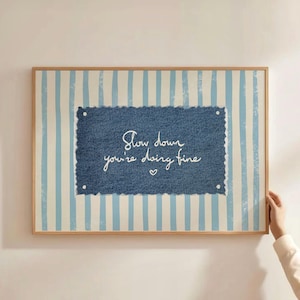 Può includere: Una stampa incorniciata con una cornice in legno chiaro. Lo sfondo presenta strisce verticali blu e bianche. Un rettangolo in denim al centro presenta il testo scritto a mano "Slow down you're doing fine" con un piccolo cuore.