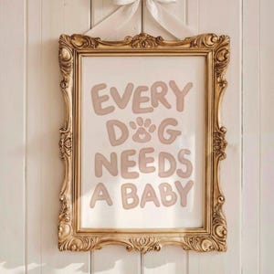 May include: A framed print with the text "EVERY D&G NEEDS A BABY" in light brown letters. The frame is ornate, gold-colored, and rectangular. A white bow is tied at the top, and the print hangs against a white paneled wall.