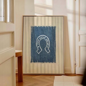 May include: Framed artwork featuring a white horseshoe design with the text "HOW LUCKY ARE WE" on a blue denim-textured background. The background has vertical beige and white stripes. The artwork is displayed in a room with a wooden floor.
