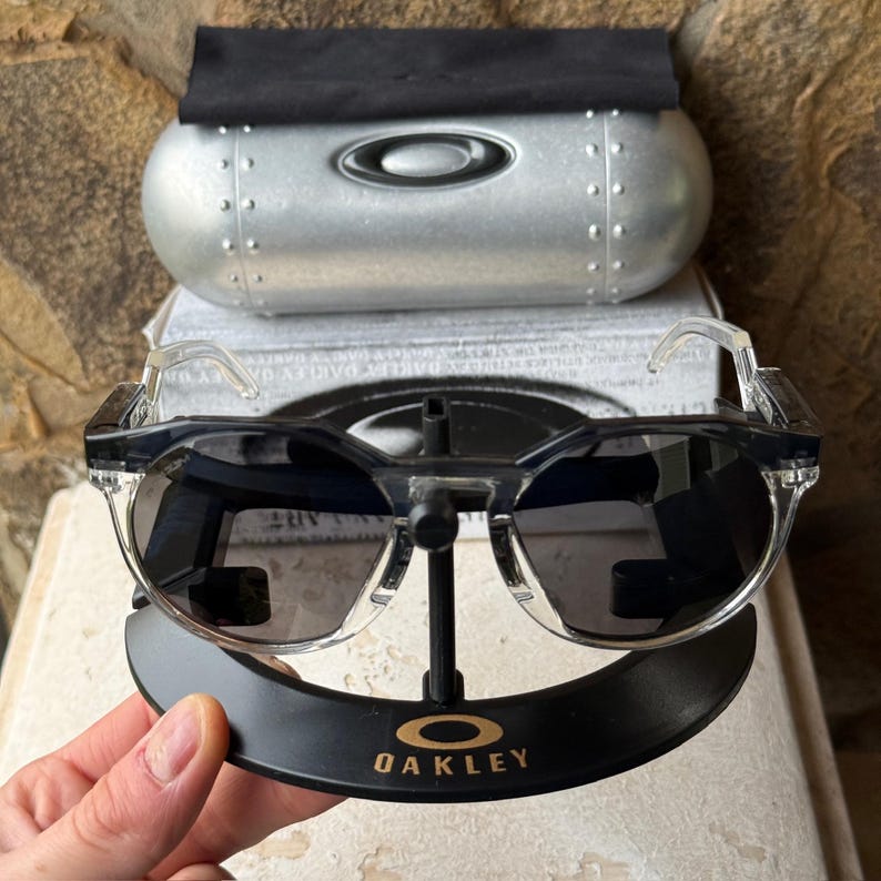May include: Clear-framed sunglasses with dark lenses are displayed on a black stand with the Oakley logo. A silver case and black cloth are behind the sunglasses. The sunglasses have a modern, sporty design.