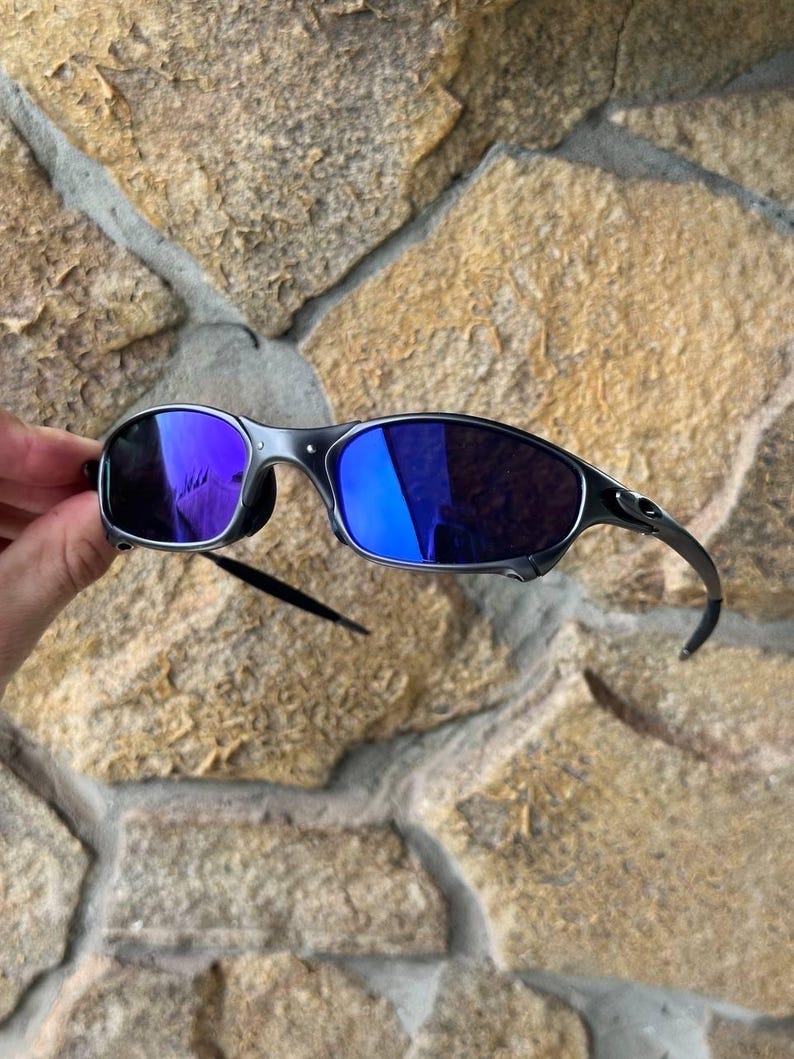 May include: A pair of sunglasses with a dark gray frame and blue mirrored lenses. The sunglasses have a sleek, modern design with curved temples and a minimalist aesthetic. The lenses reflect a vibrant blue hue.