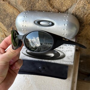 K&ouml;nnte beinhalten: Schwarze Oakley-Sonnenbrille mit gebogenem Rahmen und verspiegelten Gl&auml;sern. Die Sonnenbrille wird mit einem silbernen Etui und einem schwarzen Mikrofaserbeutel pr&auml;sentiert. Die Gl&auml;ser spiegeln eine Himmel- und Berglandschaft wider.