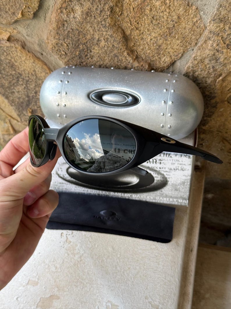 K&ouml;nnte beinhalten: Schwarze Oakley-Sonnenbrille mit gebogenem Rahmen und verspiegelten Gl&auml;sern. Die Sonnenbrille wird mit einem silbernen Etui und einem schwarzen Mikrofaserbeutel pr&auml;sentiert. Die Gl&auml;ser spiegeln eine Himmel- und Berglandschaft wider.