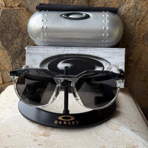 May include: Clear-framed sunglasses with dark lenses rest on a black display stand with the Oakley logo. A silver case and a box with the Oakley logo are in the background. The sunglasses have a modern, angular design.