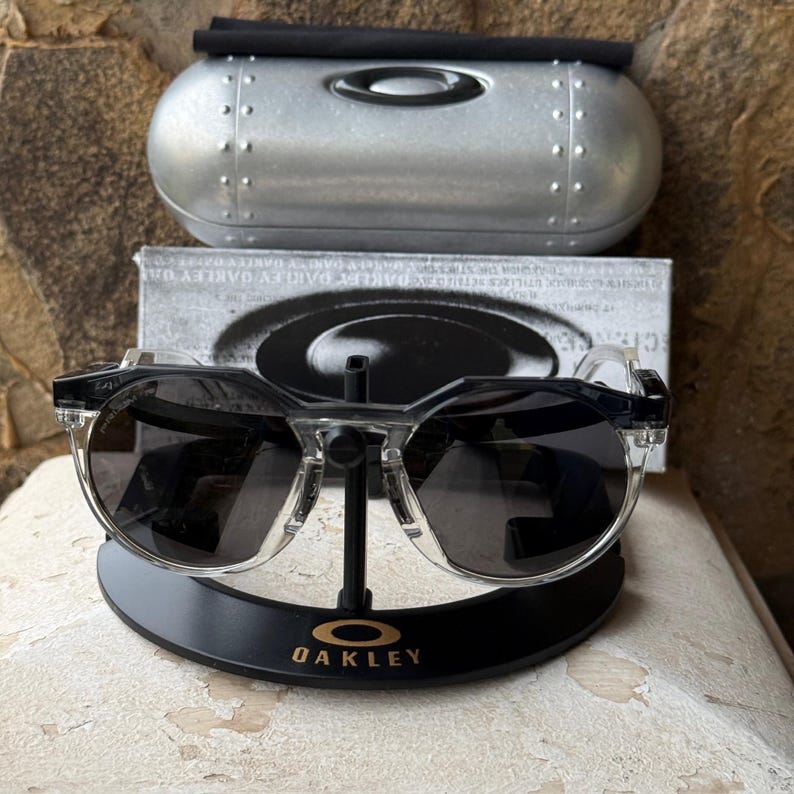 May include: Clear-framed sunglasses with dark lenses rest on a black display stand with the Oakley logo. A silver case and a box with the Oakley logo are in the background. The sunglasses have a modern, angular design.