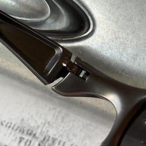 May include: Close-up of a pair of dark gray sunglasses. The sunglasses have a sleek, modern design with a curved frame and dark lenses. The hinges and arms are visible, showing the construction details.