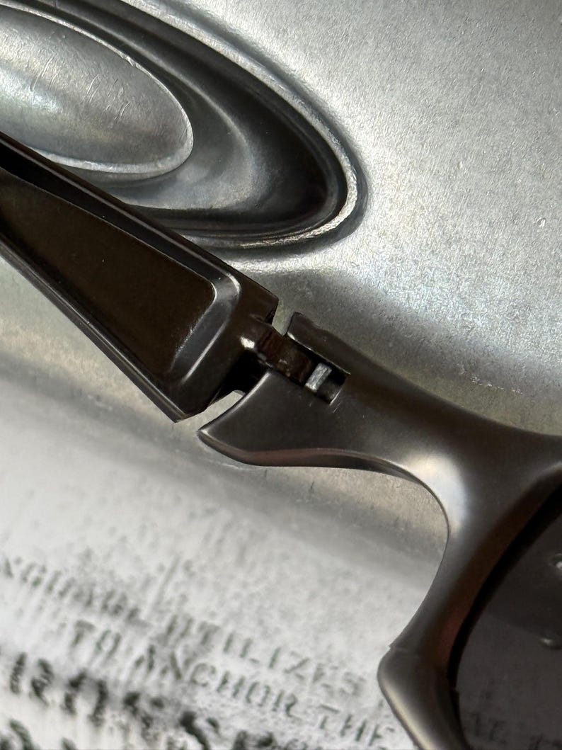 May include: Close-up of a pair of dark gray sunglasses. The sunglasses have a sleek, modern design with a curved frame and dark lenses. The hinges and arms are visible, showing the construction details.