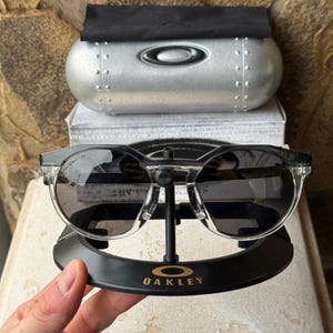 May include: Black and clear-framed sunglasses with dark lenses, displayed on a black stand with the Oakley logo. The sunglasses are paired with a silver case and a white box.