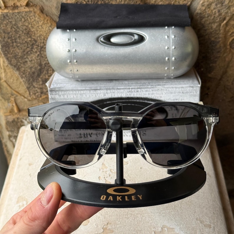 May include: Black and clear-framed sunglasses with dark lenses, displayed on a black stand with the Oakley logo. The sunglasses are paired with a silver case and a white box.