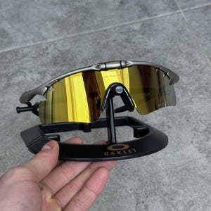 May include: Silver-framed Oakley sunglasses with gold mirrored lenses. The sunglasses are displayed on a black stand with the Oakley logo. The lenses reflect a bright yellow color.