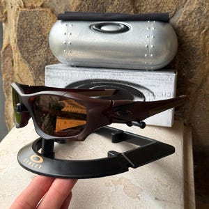 Może przedstawiać: Ciemnobrązowe okulary przeciwsłoneczne Oakley z pasującym etui i stojakiem ekspozycyjnym. Okulary przeciwsłoneczne mają elegancki, nowoczesny design z zakrzywioną oprawką i przyciemnianymi soczewkami. Etui jest srebrne z czarną g&oacute;rą.