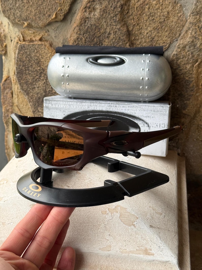 Może przedstawiać: Ciemnobrązowe okulary przeciwsłoneczne Oakley z pasującym etui i stojakiem ekspozycyjnym. Okulary przeciwsłoneczne mają elegancki, nowoczesny design z zakrzywioną oprawką i przyciemnianymi soczewkami. Etui jest srebrne z czarną g&oacute;rą.