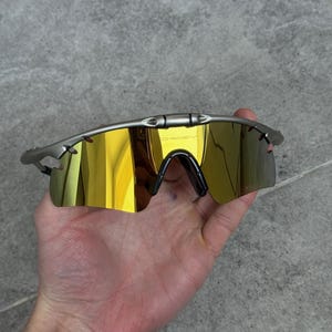 May include: Silver-framed sunglasses with gold mirrored lenses. The sunglasses have a sleek, modern design with a black nose bridge and temples. The Oakley logo is visible on the top of the frame.