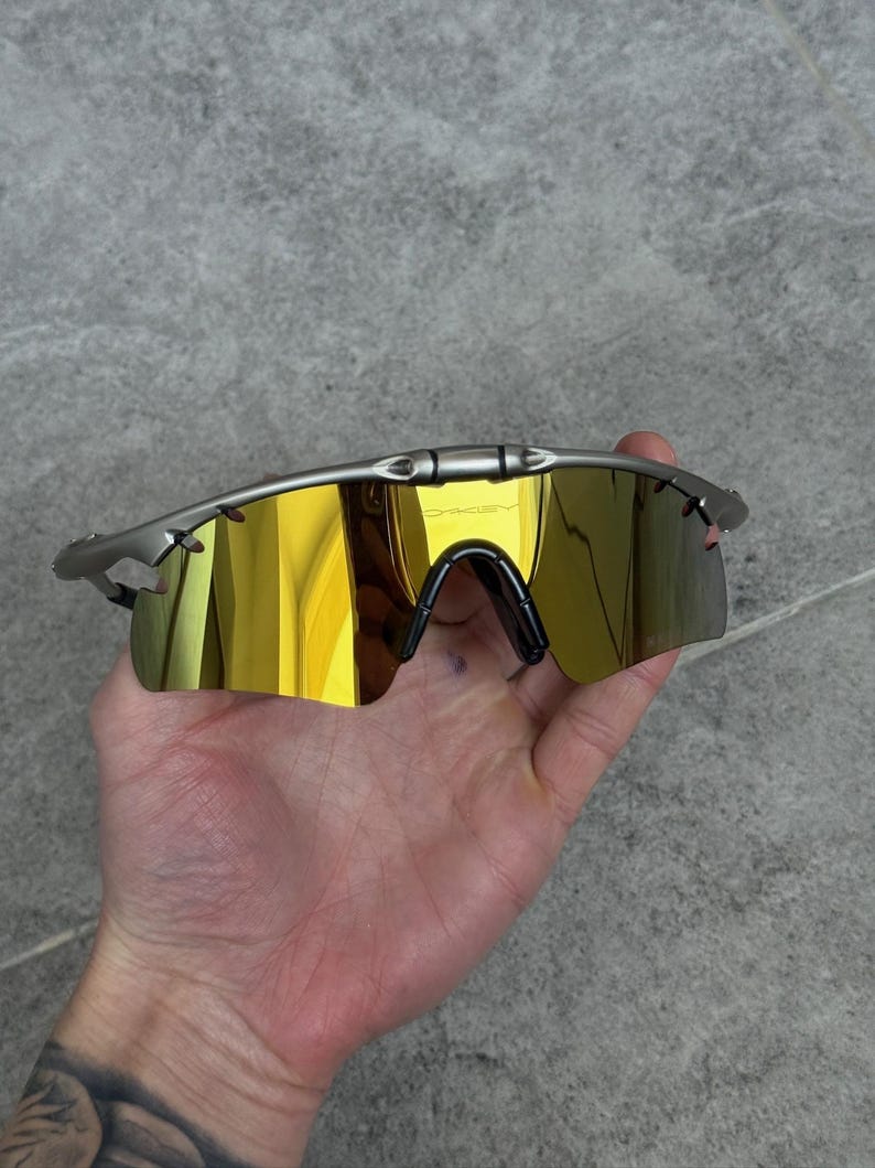 May include: Silver-framed sunglasses with gold mirrored lenses. The sunglasses have a sleek, modern design with a black nose bridge and temples. The Oakley logo is visible on the top of the frame.