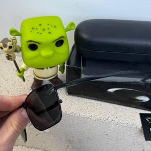 May include: Black square-framed sunglasses with dark lenses are held in front of a Shrek figurine. The sunglasses have a sleek, modern design. A black case and box are in the background.