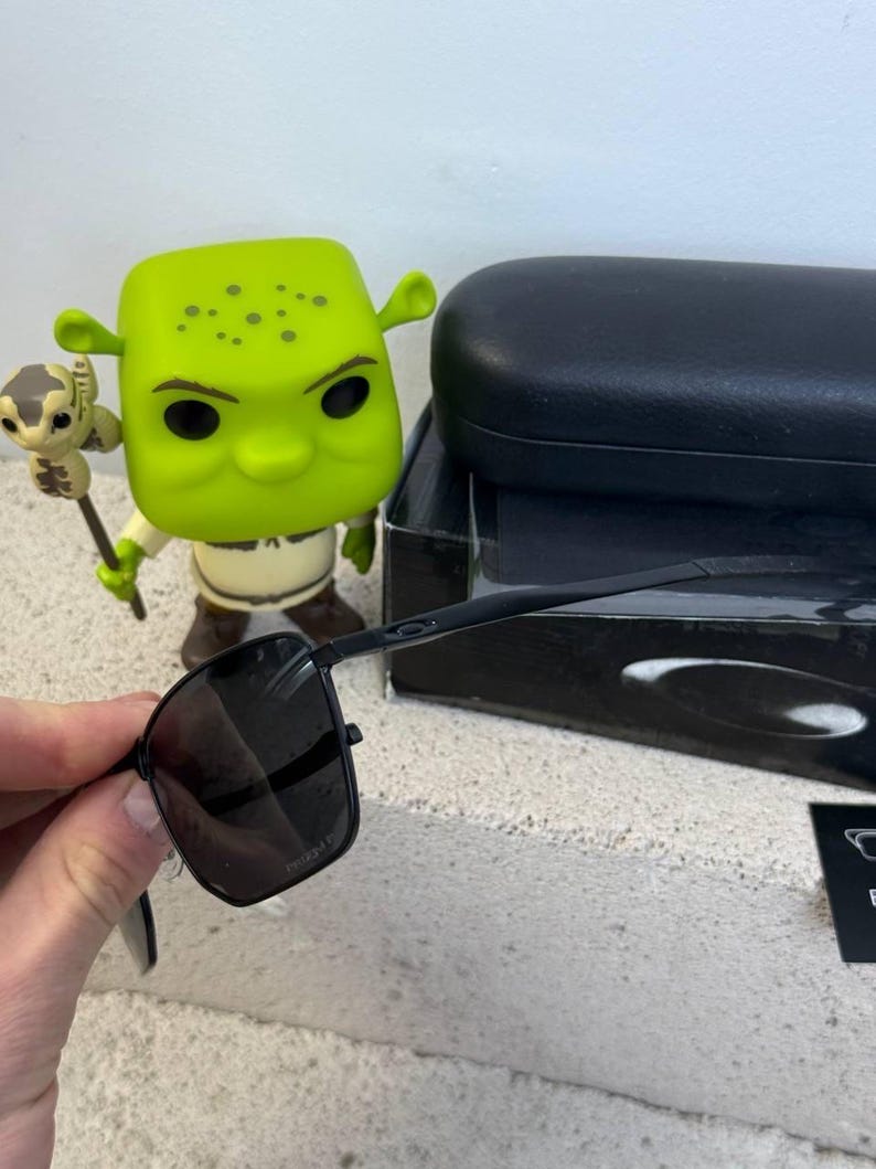 May include: Black square-framed sunglasses with dark lenses are held in front of a Shrek figurine. The sunglasses have a sleek, modern design. A black case and box are in the background.