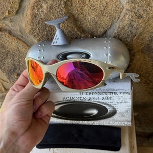 May include: Off-white Oakley sunglasses with red mirrored lenses. The glasses are held in front of a silver case and a box with the text "SCIENCE AND ART". A black case and cleaning cloth are also visible.