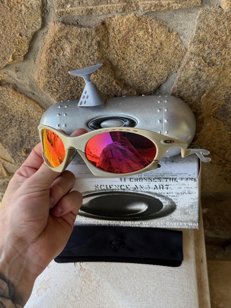 May include: Off-white Oakley sunglasses with red mirrored lenses. The glasses are held in front of a silver case and a box with the text "SCIENCE AND ART". A black case and cleaning cloth are also visible.