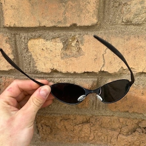 May include: Black oval sunglasses with a thin black frame. The sunglasses have dark lenses and are held against a brick wall. The sunglasses have a small silver sticker on the frame.