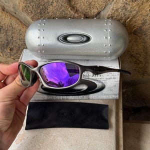 May include: Silver-framed sunglasses with purple lenses, a black case, and a black microfiber pouch. The sunglasses are displayed on top of a white box with black text. The case is silver with rivets and the Oakley logo.