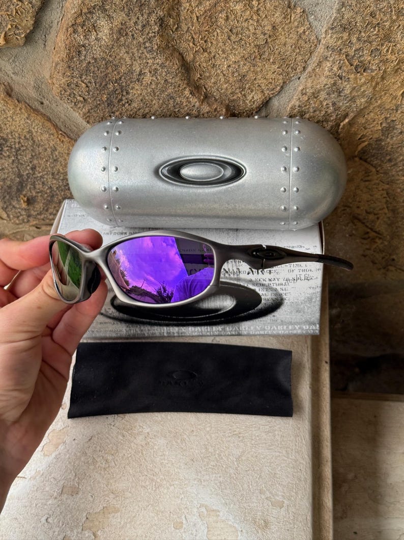May include: Silver-framed sunglasses with purple lenses, a black case, and a black microfiber pouch. The sunglasses are displayed on top of a white box with black text. The case is silver with rivets and the Oakley logo.