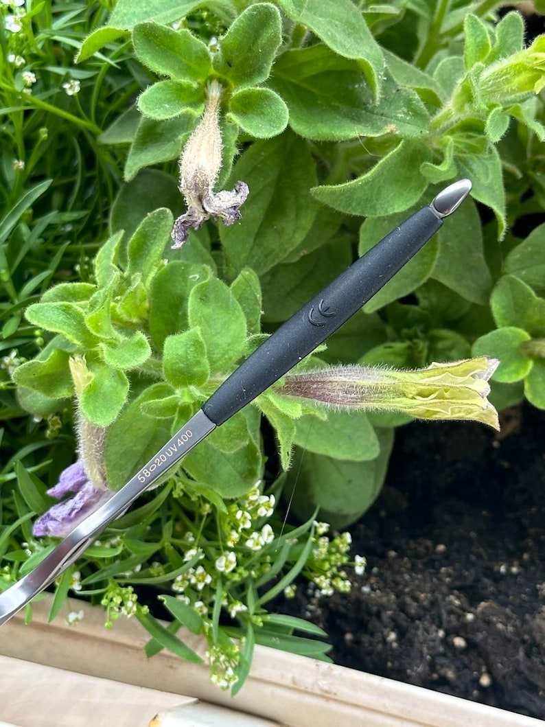 May include: Close-up of eyeglasses with a black arm and silver frame. The arm has the text "58-20 UV400". The glasses are set against a backdrop of green foliage and small white flowers.