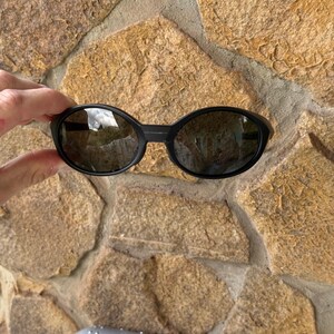 K&ouml;nnte beinhalten: Schwarze ovale Sonnenbrille mit dunklen Gl&auml;sern. Die Sonnenbrille wird vor einer strukturierten Steinmauer gehalten. Das Oakley-Logo ist auf dem silbernen Etui am unteren Bildrand zu sehen.