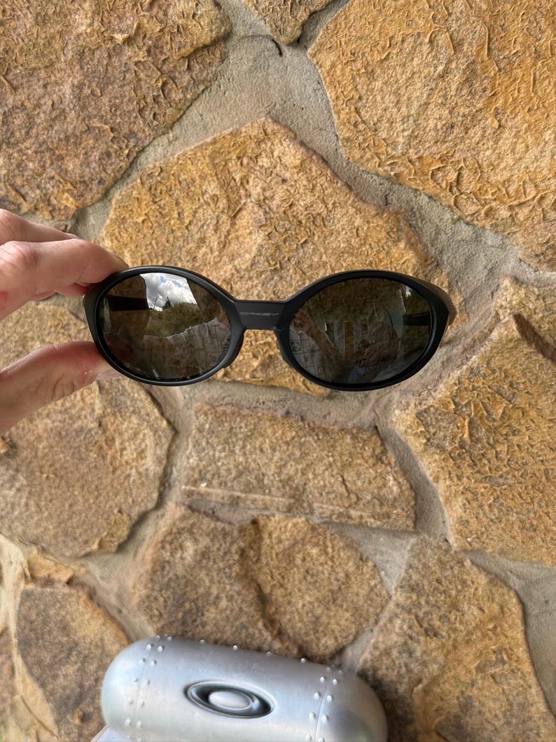 K&ouml;nnte beinhalten: Schwarze ovale Sonnenbrille mit dunklen Gl&auml;sern. Die Sonnenbrille wird vor einer strukturierten Steinmauer gehalten. Das Oakley-Logo ist auf dem silbernen Etui am unteren Bildrand zu sehen.
