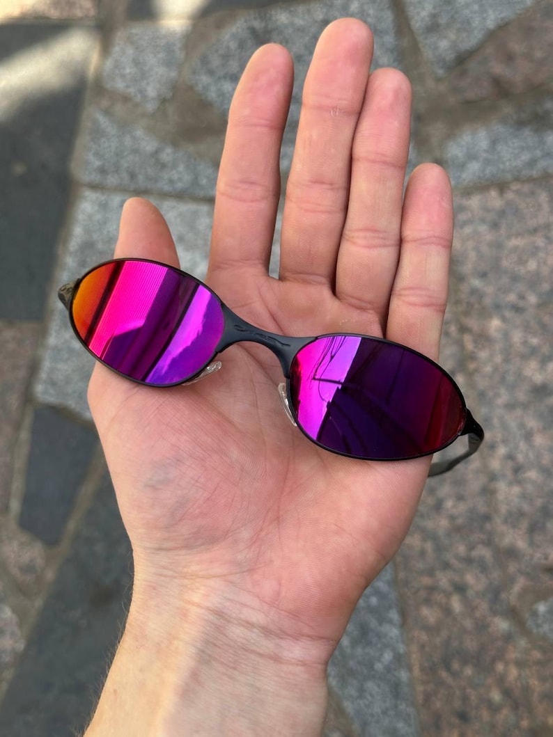 May include: Oval-shaped sunglasses with black frames and pink and purple mirrored lenses. The lenses reflect a range of colours. A contemporary eyewear accessory.